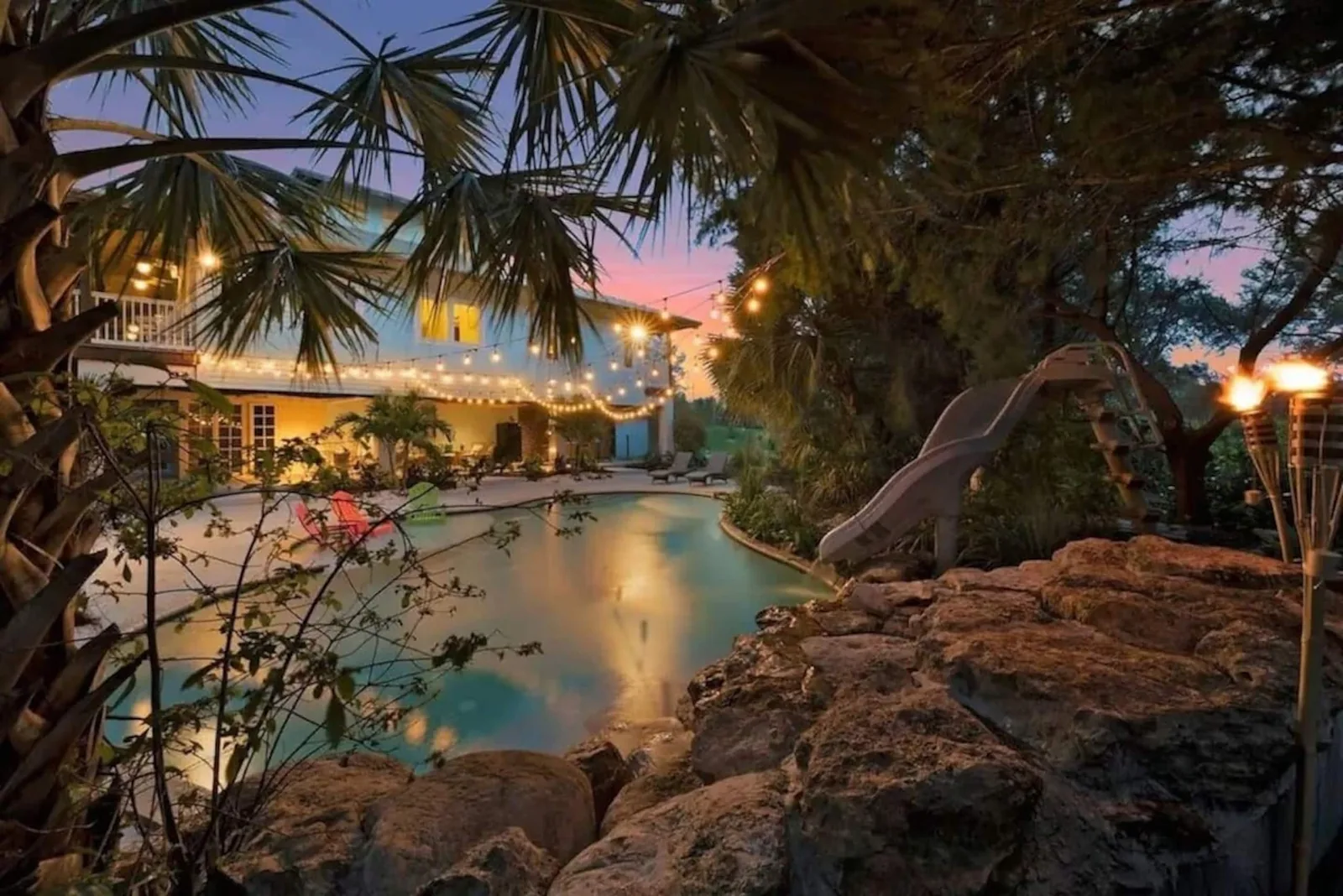 The pool at dusk seen past the rock waterfall, the house lit behind